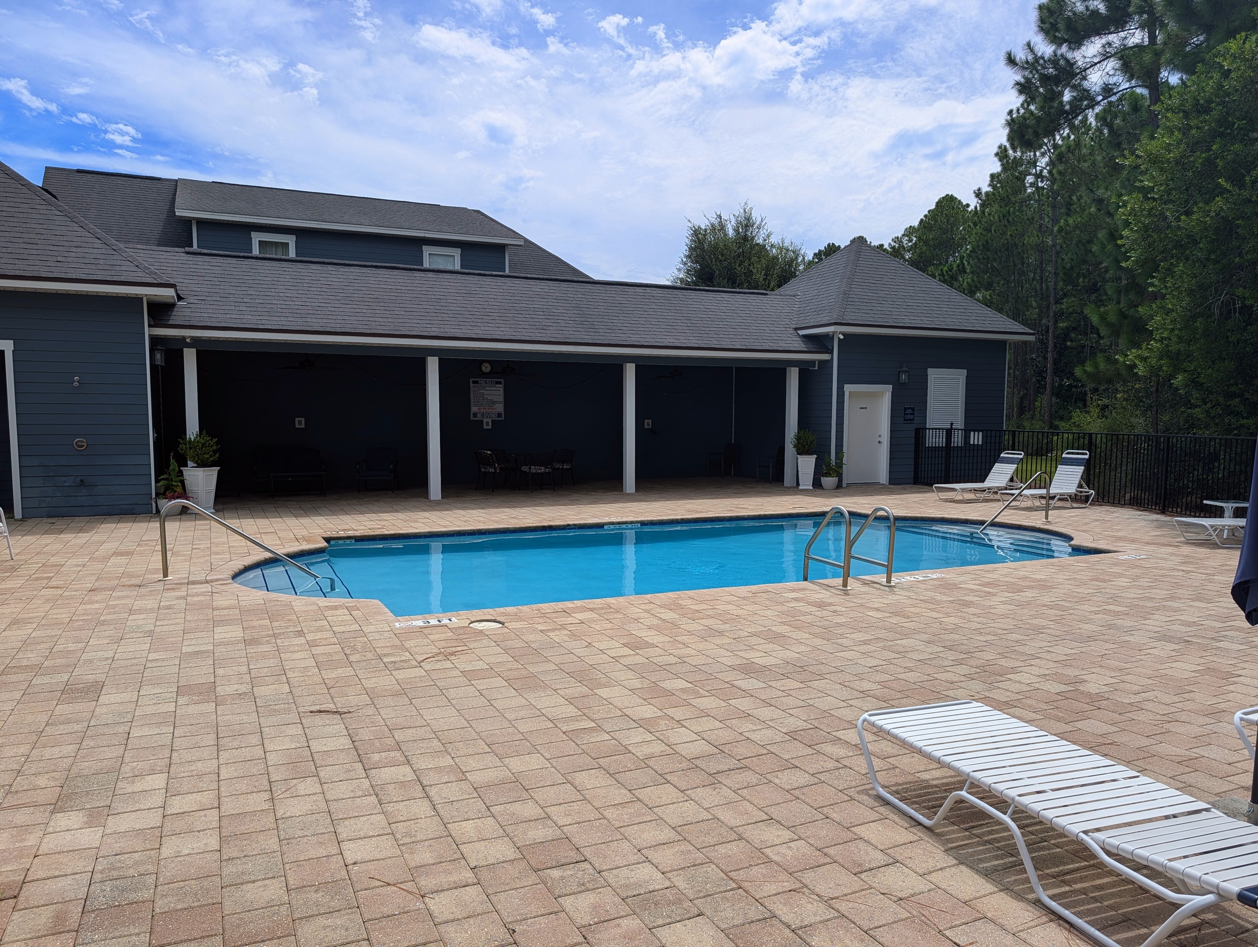 The community pool at Paradise Cove, with a brick paver deck, white loungers and a navy clubhouse with covered seating